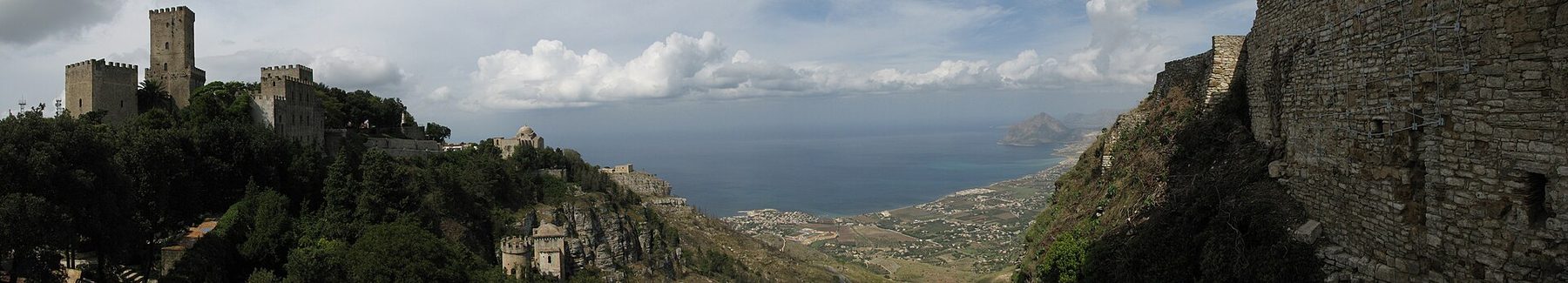 A wide panoramic view of Erice from above, showing its terracotta roofs and the curve of the Sicilian coast.